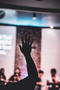 A silhouetted hand is raised in front of a blurred background where a group of people appear to be either performing or presenting. The setting looks like an indoor event with artificial lighting.