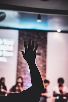 A silhouetted hand is raised in front of a blurred background where a group of people appear to be either performing or presenting. The setting looks like an indoor event with artificial lighting.