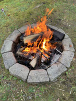 A stone-ringed fire pit is burning brightly, with flames dancing around stacked logs. The surrounding ground is covered with grass and patches of dirt, creating a rustic outdoor setting.