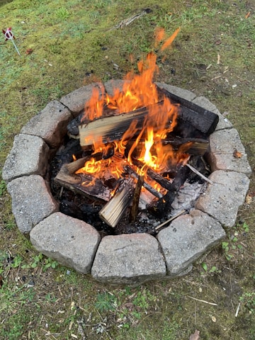 A stone-ringed fire pit is burning brightly, with flames dancing around stacked logs. The surrounding ground is covered with grass and patches of dirt, creating a rustic outdoor setting.