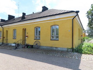 A yellow stone building with a gabled black metal roof and small chimneys. There are arched windows with white trim and a few potted flowers near the entrance. A wooden wagon wheel rests against the wall. Cobblestone and gravel paths surround the building, with green bushes and trees in the background.