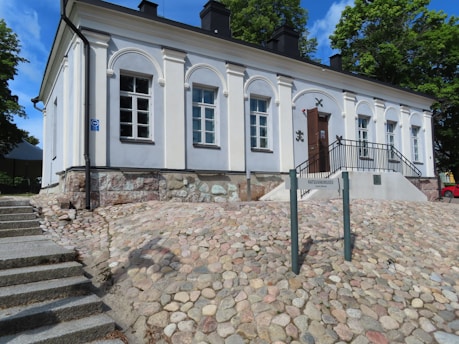A historic building with multiple large windows and a facade featuring arch designs and pillars. The structure is situated on a stone-paved slope with stairs to the side. A dark door with metal decorations is accessible via a small metal staircase with a railing. Mature trees and a blue sky form the background.