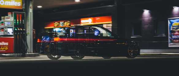 A car is parked at a gas station at night, with visible motion blur creating a ghostly effect. The station is well-lit with several glowing signs and advertisements. The environment appears deserted, adding a sense of solitude.