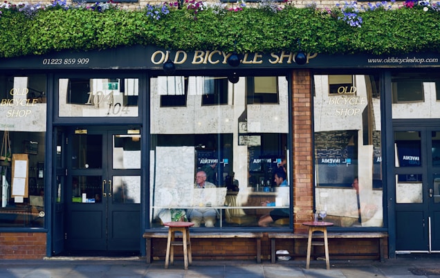 A quaint storefront featuring a vintage-style sign reading 'Old Bicycle Shop' with green plants and flowers adorning the awning. The shop has large windows with reflections of the street and inside seating. Patrons can be seen sitting at tables just inside the window.