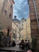 An outdoor cafe located in a narrow, historic alley with old, weathered buildings and graffiti-covered walls. Several people are seated at tables, enjoying drinks and conversation. In the background, there are domed architecture and a cloudy sky, enhancing the vintage ambiance.