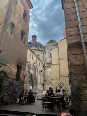 An outdoor cafe located in a narrow, historic alley with old, weathered buildings and graffiti-covered walls. Several people are seated at tables, enjoying drinks and conversation. In the background, there are domed architecture and a cloudy sky, enhancing the vintage ambiance.