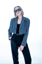 A person with short, light brown hair wears oversized dark sunglasses and a stylish outfit consisting of a blue-grey blazer over a black top and black trousers. The person has one hand in their pocket and exudes a confident demeanor against a bright, minimalist background.