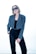 A person with short, light brown hair wears oversized dark sunglasses and a stylish outfit consisting of a blue-grey blazer over a black top and black trousers. The person has one hand in their pocket and exudes a confident demeanor against a bright, minimalist background.