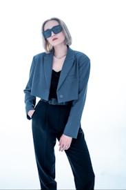 A person with short, light brown hair wears oversized dark sunglasses and a stylish outfit consisting of a blue-grey blazer over a black top and black trousers. The person has one hand in their pocket and exudes a confident demeanor against a bright, minimalist background.