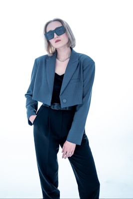 A person with short, light brown hair wears oversized dark sunglasses and a stylish outfit consisting of a blue-grey blazer over a black top and black trousers. The person has one hand in their pocket and exudes a confident demeanor against a bright, minimalist background.