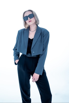 A person with short, light brown hair wears oversized dark sunglasses and a stylish outfit consisting of a blue-grey blazer over a black top and black trousers. The person has one hand in their pocket and exudes a confident demeanor against a bright, minimalist background.