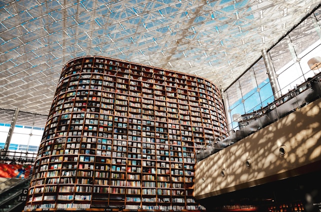 A large, modern library with an impressive, curved bookshelf that extends from the floor to the ceiling is filled with a vast array of books. The ceiling is made of a geometric, glass-like material allowing natural light to flood into the open space. Shadows cast intricate patterns on the walls and floor, creating an airy and sophisticated atmosphere.