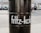 A black barrel with the name 'fritz-kola' written in white letters. It is standing on a paved surface with several parked cars and a few people in the background. The environment appears to be urban, possibly a parking lot or street.
