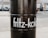 A black barrel with the name 'fritz-kola' written in white letters. It is standing on a paved surface with several parked cars and a few people in the background. The environment appears to be urban, possibly a parking lot or street.