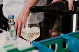 A person is pouring a clear liquid, likely wine, into a wine glass. There is a bottle of mineral water placed on a table next to the glass, and blue plastic crates are partially visible in the foreground.