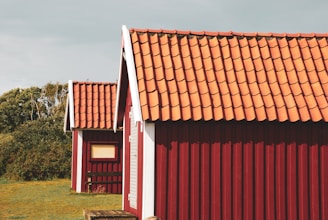 Two rustic red-painted wooden buildings with white trim and clay-tiled roofs are situated on a grassy area. Dense foliage is visible in the background under a slightly overcast sky.
