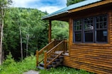 A wooden cabin with a green roof is situated in a lush forest setting. The cabin has large dark-framed windows and a small set of steps leading to a porch. Surrounding the cabin are dense green trees and a well-maintained grassy area under a partly cloudy sky.
