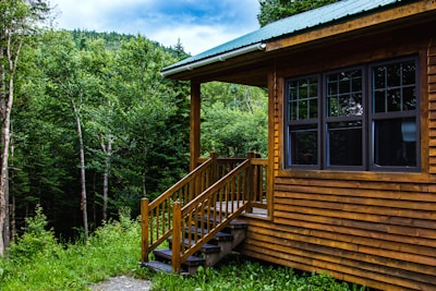 A wooden cabin with a green roof is situated in a lush forest setting. The cabin has large dark-framed windows and a small set of steps leading to a porch. Surrounding the cabin are dense green trees and a well-maintained grassy area under a partly cloudy sky.
