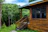 A wooden cabin with a green roof is situated in a lush forest setting. The cabin has large dark-framed windows and a small set of steps leading to a porch. Surrounding the cabin are dense green trees and a well-maintained grassy area under a partly cloudy sky.