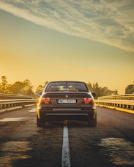 A black BMW with the license plate 'ML-5951' is parked on an empty road during sunrise or sunset. The sky is filled with warm hues of yellow and orange, creating a serene atmosphere. The perspective is from behind the car, emphasizing the road stretching ahead.