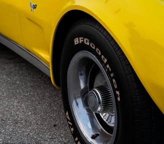 A close-up view of a classic car tire, featuring polished chrome rims and a BFGoodrich tire with visible lettering. The tire is mounted on a bright yellow vehicle, partially visible, with a reflective surface. The ground is made of asphalt.