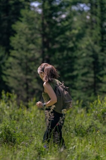 A person is hiking through a lush, green forest. They are wearing a backpack and a sleeveless top, suggesting a casual outdoor exploration. The surroundings are filled with tall trees and dense vegetation, indicating a rich, natural environment.