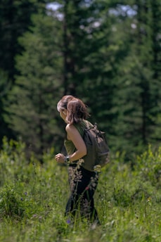 A person is hiking through a lush, green forest. They are wearing a backpack and a sleeveless top, suggesting a casual outdoor exploration. The surroundings are filled with tall trees and dense vegetation, indicating a rich, natural environment.