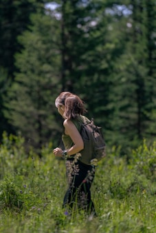 A person is hiking through a lush, green forest. They are wearing a backpack and a sleeveless top, suggesting a casual outdoor exploration. The surroundings are filled with tall trees and dense vegetation, indicating a rich, natural environment.