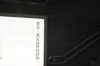 A wall-mounted illuminated advertisement display with Chinese text on the left side against a dark wall. A staircase is visible on the right with a metal handrail.