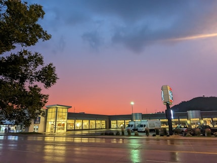 A commercial building with large glass windows is illuminated against a stunning sunset backdrop featuring vibrant pink and blue hues. A wet street in the foreground reflects the lights from the building and street lamps. A tree is visible on the left, and a hill can be seen in the background. There is a signpost in front of the building and vehicles parked nearby.
