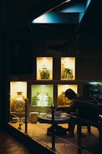 A dimly illuminated display showcases various intricately decorated ceramic vases and pottery pieces. The warm light highlights a figure at a pottery wheel, adding depth to the exhibit of artistic craftsmanship.