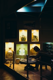 A dimly illuminated display showcases various intricately decorated ceramic vases and pottery pieces. The warm light highlights a figure at a pottery wheel, adding depth to the exhibit of artistic craftsmanship.