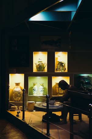 A dimly illuminated display showcases various intricately decorated ceramic vases and pottery pieces. The warm light highlights a figure at a pottery wheel, adding depth to the exhibit of artistic craftsmanship.