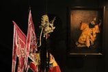 A detailed and ornate traditional costume stands in the foreground, featuring elaborate embroidery and vibrant colors such as reds and golds, with a distinctive headdress possibly used in a cultural ceremony or performance. In the background, a painting portrays a character with a dramatic pose, wearing a costume that includes a long beard and similarly extravagant attire, suggesting a connection to traditional theatrical performances.