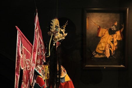 A detailed and ornate traditional costume stands in the foreground, featuring elaborate embroidery and vibrant colors such as reds and golds, with a distinctive headdress possibly used in a cultural ceremony or performance. In the background, a painting portrays a character with a dramatic pose, wearing a costume that includes a long beard and similarly extravagant attire, suggesting a connection to traditional theatrical performances.