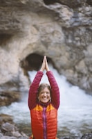 A woman in a colorful jacket stands in front of a rocky waterfall with her hands raised and pressed together above her head. She is smiling broadly, appearing cheerful and energetic.