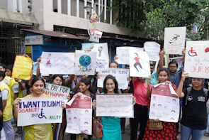 A diverse group of people are gathered for a protest or awareness event, holding various colorful signs advocating for disability rights and inclusion. Some signs have text promoting the inclusion of individuals with disabilities in sports and society, with messages like 'I want to turn my disability into ability' and 'Support Disability Rights.' The participants appear engaged and passionate about the cause.