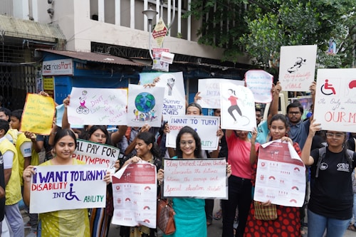 A diverse group of people are gathered for a protest or awareness event, holding various colorful signs advocating for disability rights and inclusion. Some signs have text promoting the inclusion of individuals with disabilities in sports and society, with messages like 