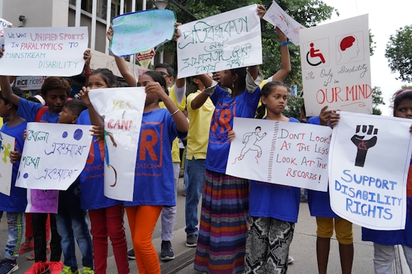 A group of children and adults are holding up various signs advocating for disability rights and inclusion in sports. The signs have messages like 'We want unified sports in the Paralympics to mainstream disability,' 'Support disability rights,' 'Don't look at the less, look at the records,' and 'Yes I can.' The participants are wearing blue shirts with orange and red lettering and are standing on an outdoor street with trees and buildings in the background.