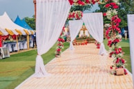 A decorative outdoor setting with a long aisle covered by a white, sheer canopy adorned with vibrant red flowers. On either side of the aisle are decorative floral arrangements and greenery. In the background, tented structures with colorful drapes provide a festive atmosphere, and a few people are visible near the tents.