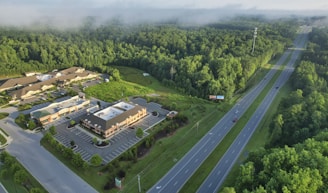 An aerial view of a commercial area surrounded by dense forest. The image shows a series of buildings with parking lots situated near a wide highway with a few cars. The landscape is dominated by lush green trees, and mist is visible above the forest.