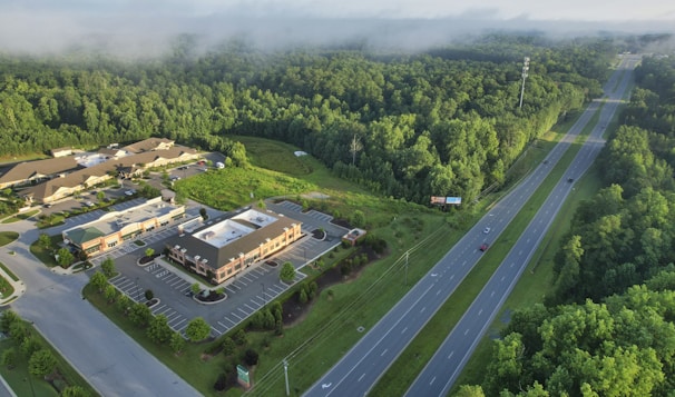 An aerial view of a commercial area surrounded by dense forest. The image shows a series of buildings with parking lots situated near a wide highway with a few cars. The landscape is dominated by lush green trees, and mist is visible above the forest.