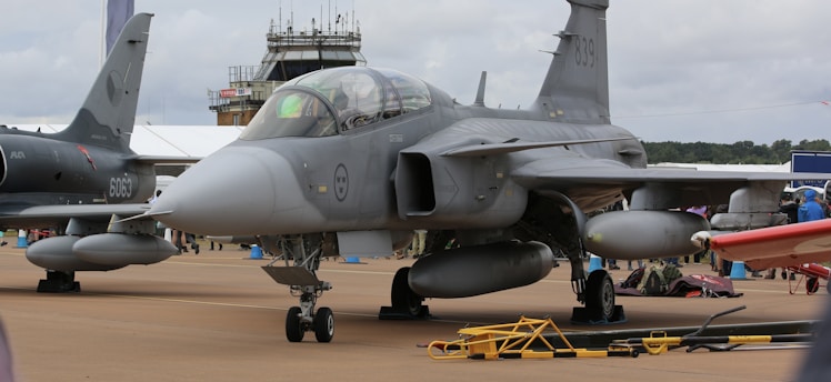 A fighter jet is parked on an airfield with cloudy skies above. The aircraft is gray and features various military insignias. In the background, a control tower is visible alongside another jet.