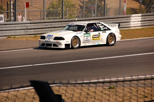 A white race car is driving on a race track with various sponsor logos on its body. The car features gold-colored rims and a rear spoiler. There is a chain-link fence on the right and a protective barrier lining the track. Sunlight casts shadows on the asphalt.