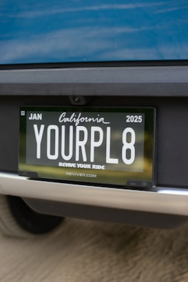 Close-up view of a car's rear license plate with a custom plate that reads 'YOURPL8'. The license plate is issued from California, valid until January 2025. The car's paint appears to be blue.