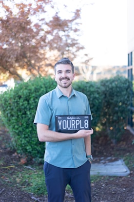 A smiling man stands outdoors holding a black license plate with the text 'YOURPL8'. He wears a light blue shirt and dark pants. The background features lush green bushes and a tree with autumn-colored leaves.