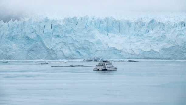 A large glacier towers over a body of water with a tourist boat in the foreground. The glacier is vast, rugged, and has a light blue hue. The sky is overcast, adding to the cold, icy atmosphere.