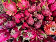 A cluster of vibrant dragon fruits with their distinct reddish-pink skins and green scaly leaves. The fruits are piled closely together, displaying various stages of ripeness, from bright pink to a deeper red. Some fruits show natural blemishes and textured surfaces, adding to the organic feel.