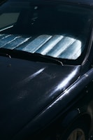 A close-up view of a car windshield with a reflective sunshade in place to block sunlight. The surface of the car is visible with reflections and subtle scratches on a dark paint.