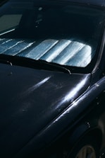 A close-up view of a car windshield with a reflective sunshade in place to block sunlight. The surface of the car is visible with reflections and subtle scratches on a dark paint.